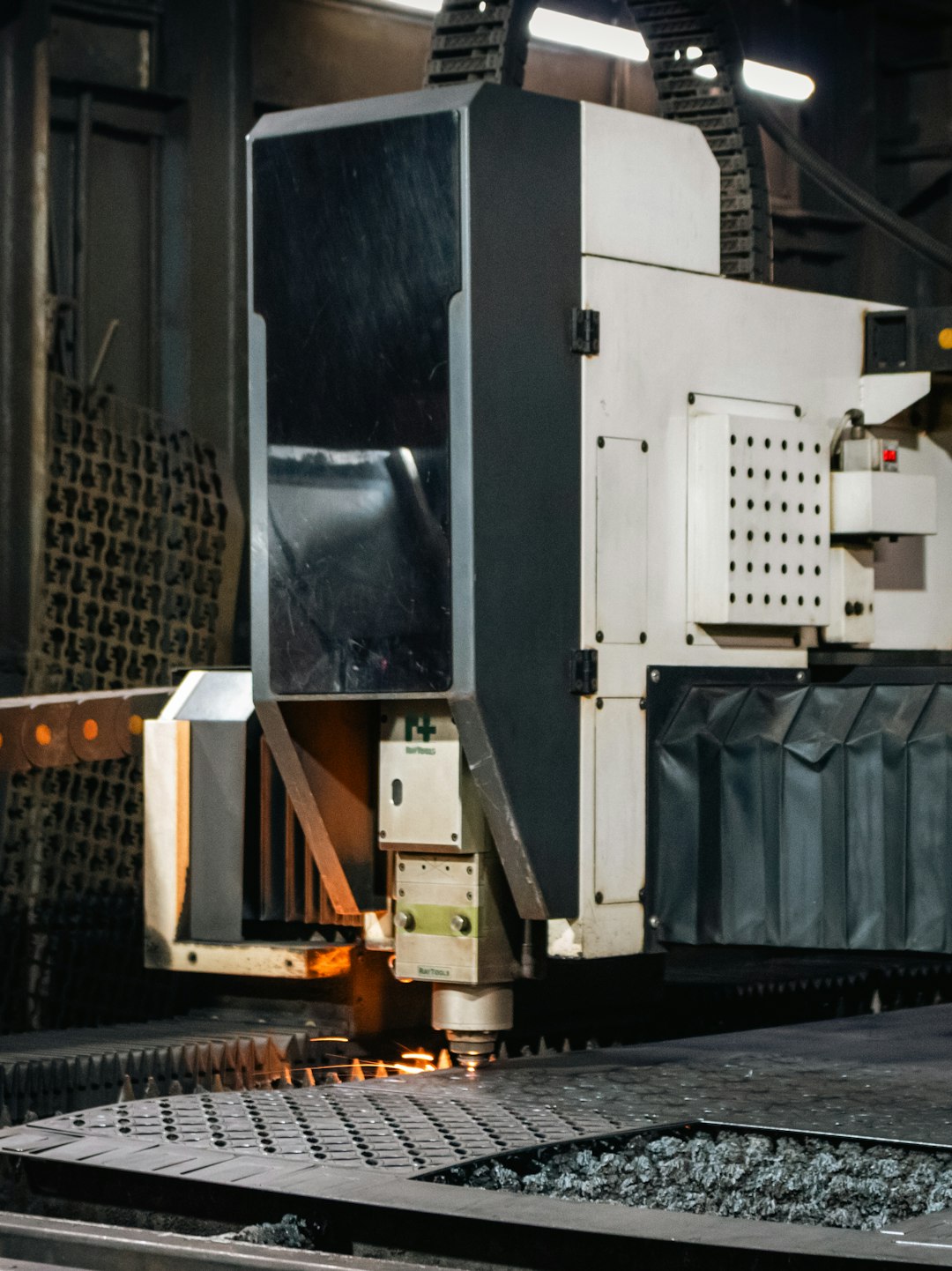 About A close-up captures the action of an industrial CNC laser/plasma machine actively cutting a metal sheet. The cutting head is visible, emitting a small spark or glow of heat as it moves across the metal surface supported by a spiked cutting bed.